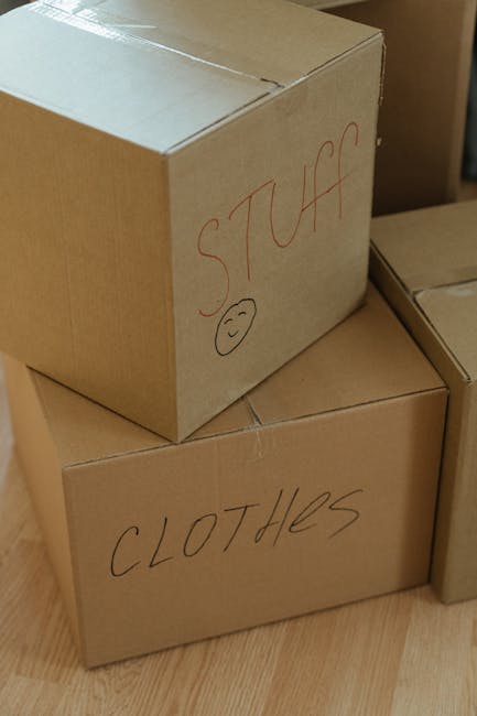Close-up view of three cardboard boxes stacked indoors, with the top box labeled 'STUFF' in red marker and a smiley face drawn underneath, and the bottom box labeled 'CLOTHES' also marked with red mar